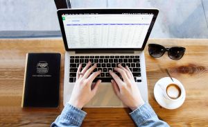 Osoba pracująca na laptopie. Obok laptopa znajdują się kawa, okulary i notatnik.