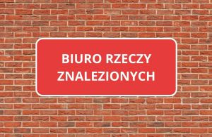 Czerwona tablica z napisem BIURO RZECZY ZNALEZIONYCH na ceglanym budynku.