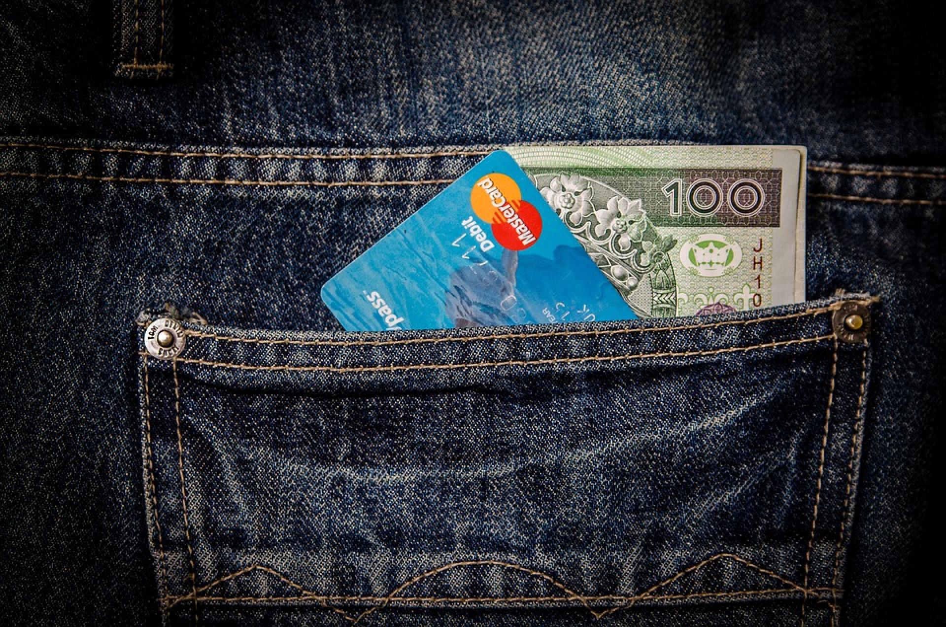 Banknot 100 złotych i karta płatnicza wystające z kieszeni dżinsowych spodni